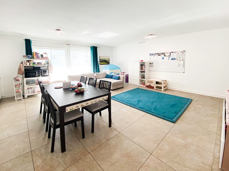 Dining table with chairs, white walls, beige floor, blue rug, white microwave, bookshelves, whiteboard, children's toys