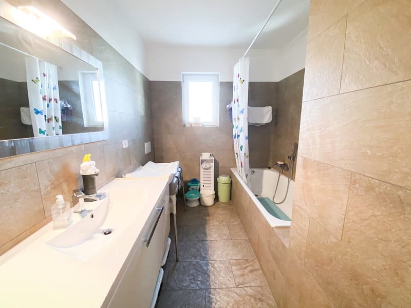 Double sink with cabinets, separate shower cubicle, bathtub, floor and wall tiles, natural light, towel rack, and various bathroom accessories.