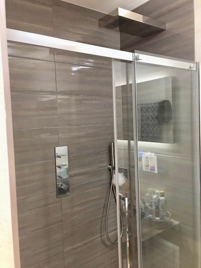 Modern bathroom, glass shower enclosure, silver shower head, tiled wall, silver faucet, shelf with personal care items, mirror with reflection of towel.