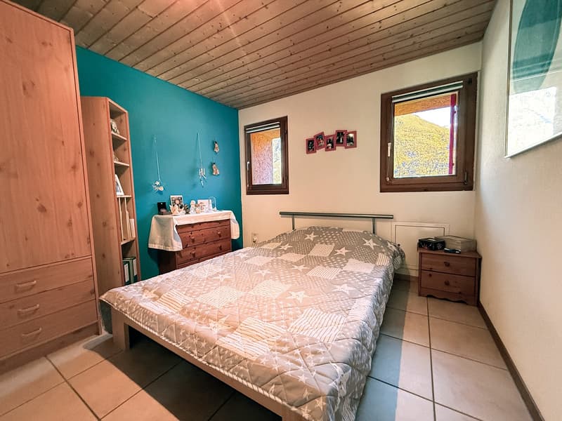 Blue walls, wooden floor, a bed with quilt, a nightstand, and a wooden cabinet, two windows, a wooden wardrobe.