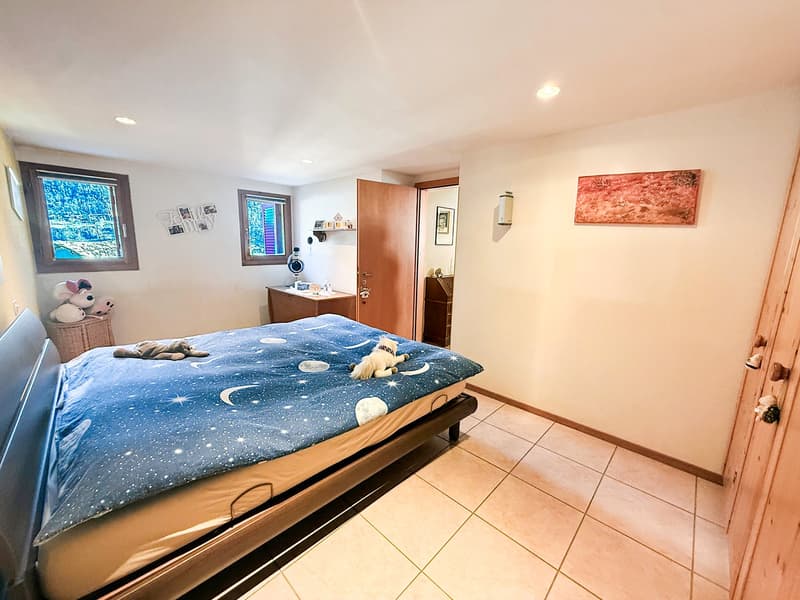Bedroom with bed, two small windows, beige tiled floor, wooden headboard, a desk with drawers, and a rack with stuffed toys