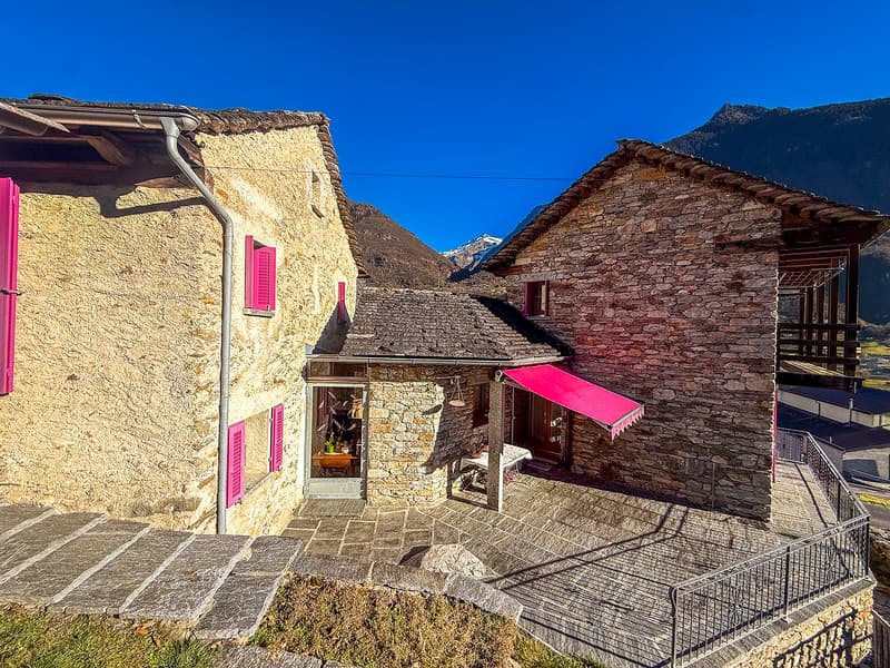 Stone building with red awning, pink shutters, stairs, metal railing, grassy slope, distant mountains