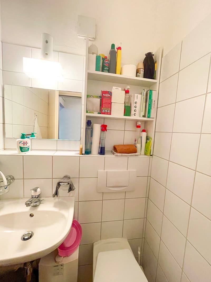 tiled walls, mirror, sink, toilet, shelves with various cleaning products, small basket, power outlet, white fixtures