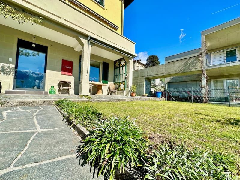 A well-maintained building with a stone pathway, garden, and plants. In front, there is a bench and a potted plant near the entrance, along with a red board. A metal fence is present. Next to the house, there is another building, with a balcony and a glas