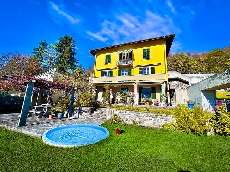 Large yellow house, wooden balcony, well-kept garden, pool, outdoor dining area, stone steps, lush greenery