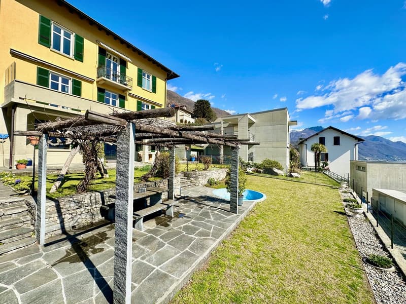 Two-story yellow building with green shutters, small swimming pool, wooden pergola, table and bench, fenced garden, gravel path