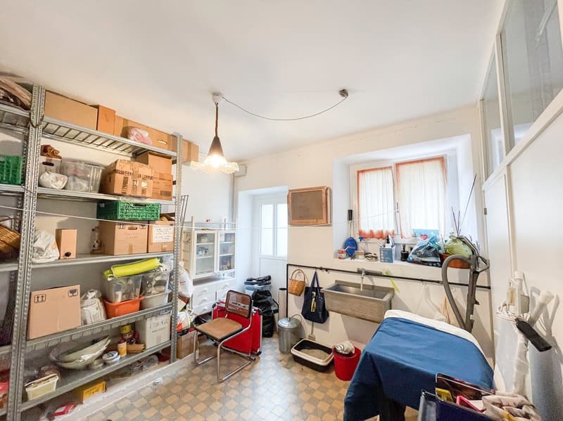 The room has a tiled floor, a bed covered with a blue sheet, shelves with storage boxes, a chair, a sink, various items, a cabinet, and lamps. There is a basket and a storage box on the floor near the cabinet.