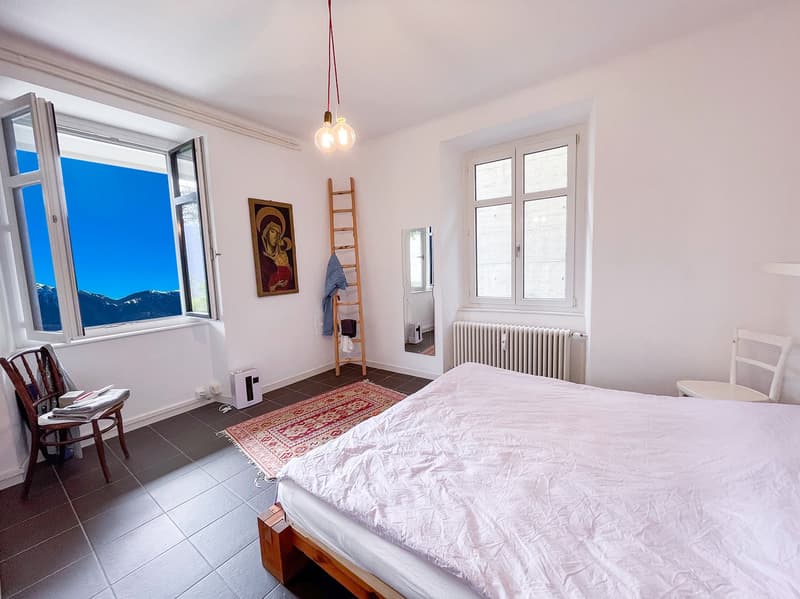 wooden bed, white bedding, white walls, white ceiling, oriental rug, large windows with mountain view, small wooden chair, small white radiator, white chair, wooden ladder, picture on wall