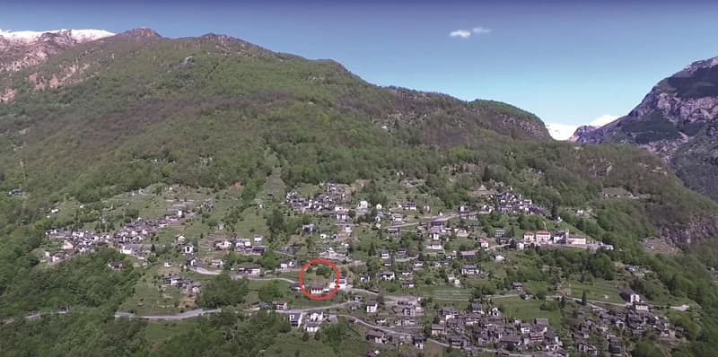 Mountain village, surrounded by hills and mountains, with houses built close together, trees and greenery all around, a large red circle highlighting a specific house