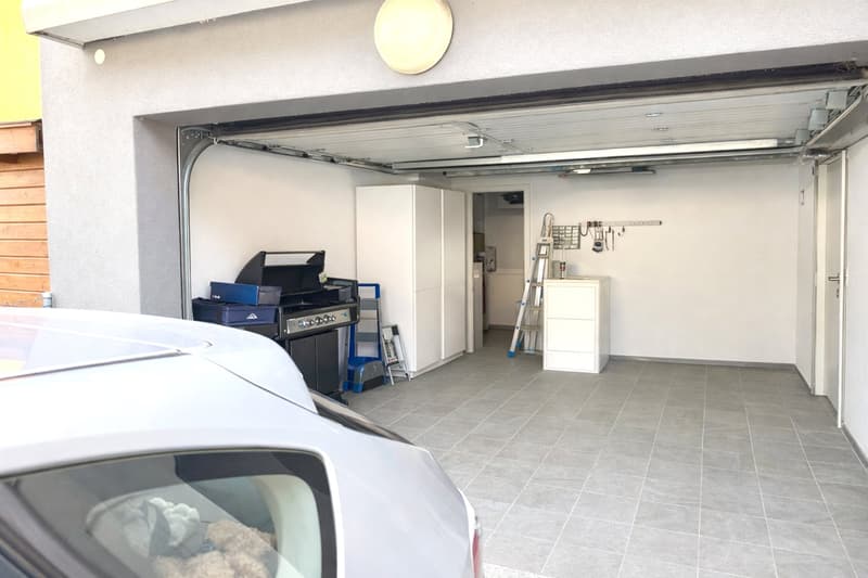 Clean, empty garage with a white car parked inside, white storage cabinets, a ladder, and tools mounted on the wall