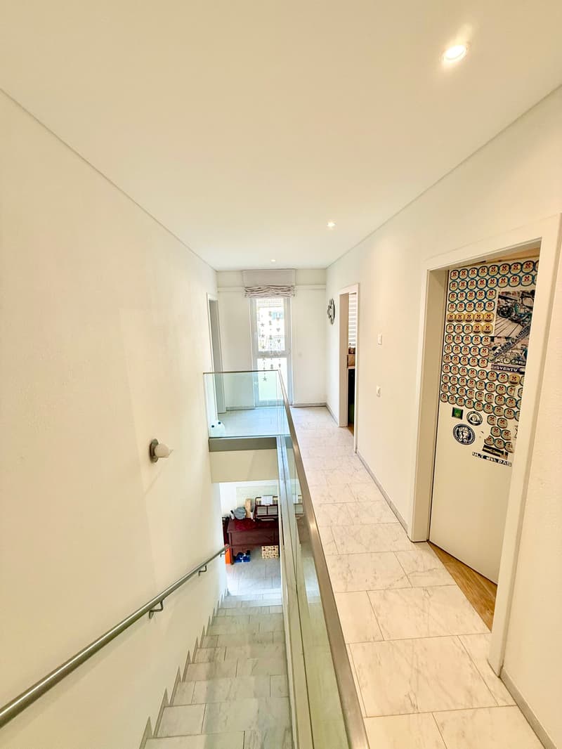 A well lit hallway with a marble staircase and glass railings, and a door at the end.