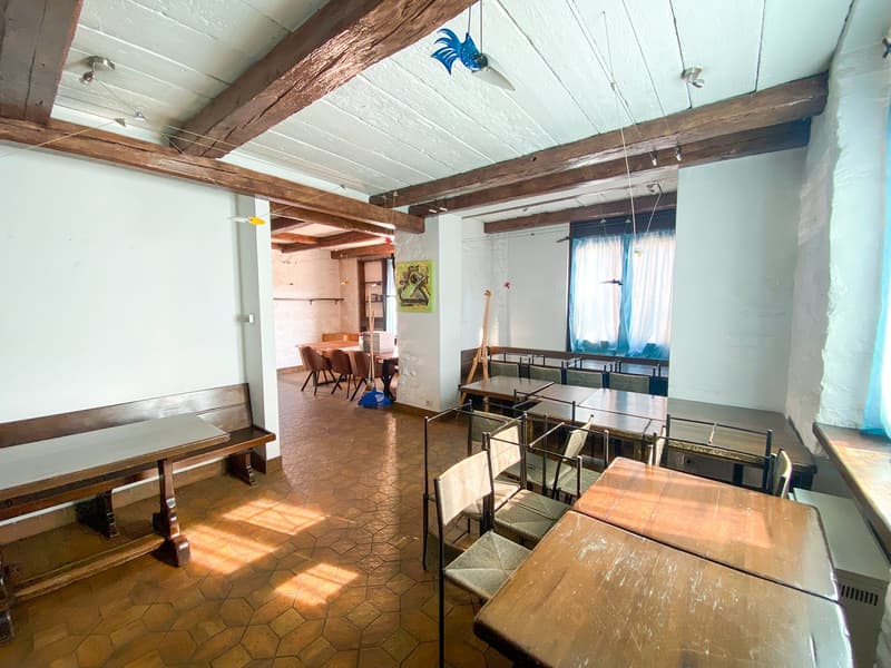 Wooden ceiling beams, wooden benches, tiled floor, long wooden tables, windows with white curtains, framed artwork on the wall.