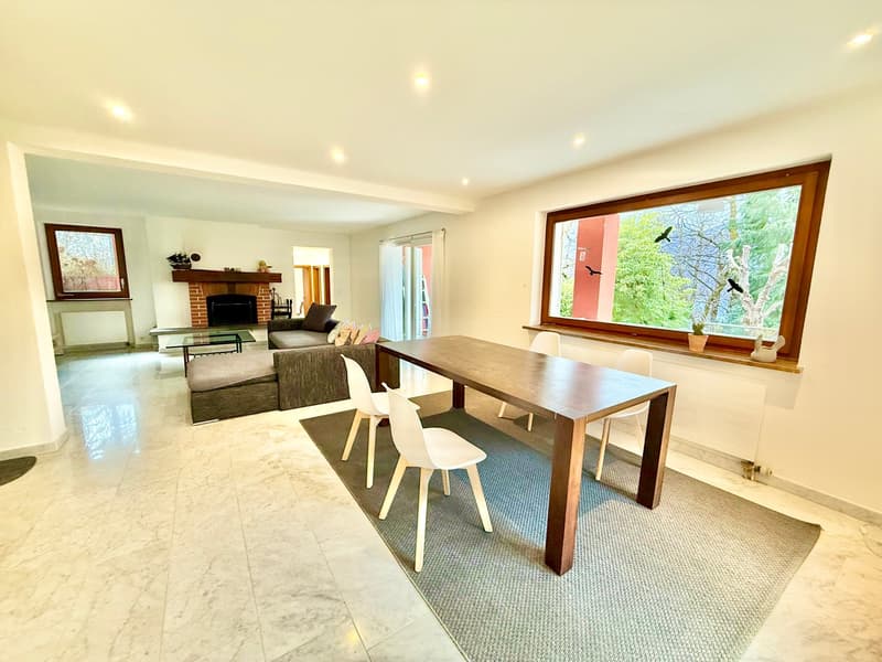 Large open room with a marble floor, a white dining table, and white chairs with a grey rug. It has a brick fireplace with a wood mantle and a large picture window.