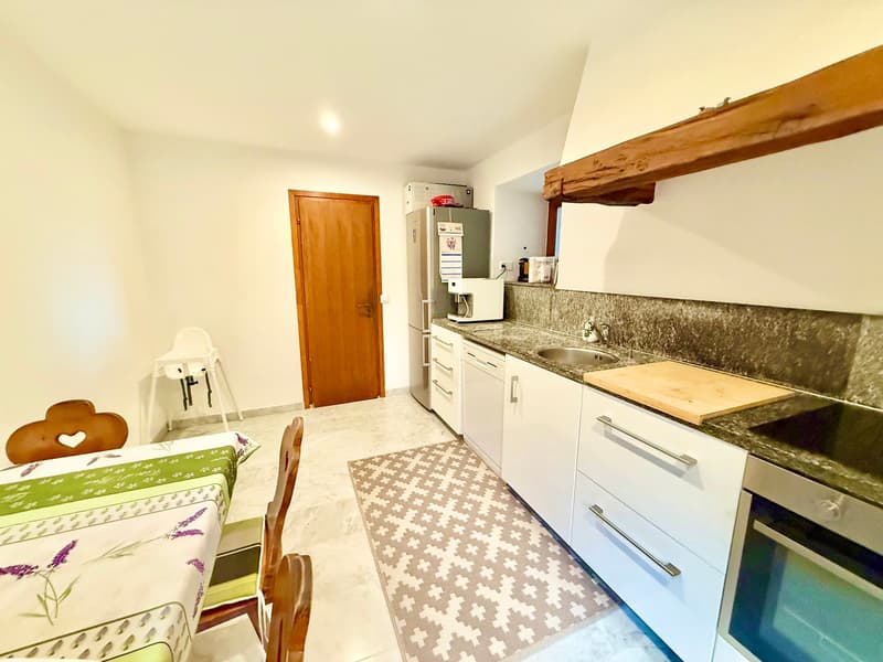 Kitchen with tiled floor, white cabinets, granite countertops, stainless steel appliances, a wooden beam, a wooden door, a rug, and a dining table with a flowered tablecloth.