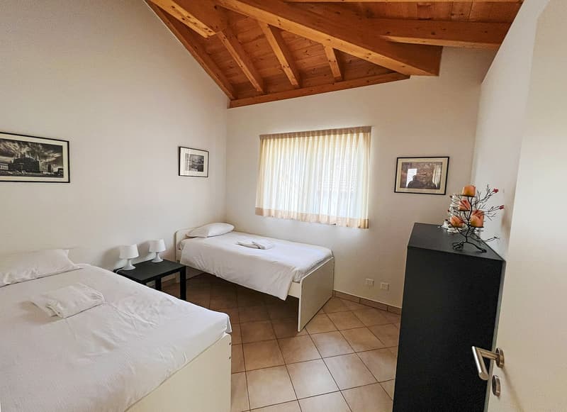 The image shows a bedroom with a vaulted wooden ceiling, two twin beds, a dresser, and framed artwork on the walls. The room has a cozy and rustic feel with the exposed wooden beams and tile flooring.