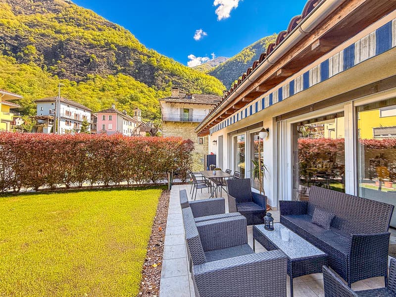 Outdoor seating area with wicker furniture, surrounded by lush greenery and mountains in the background. The area has a paved walkway and a grassy lawn. The building has a wooden roof and colorful facades.