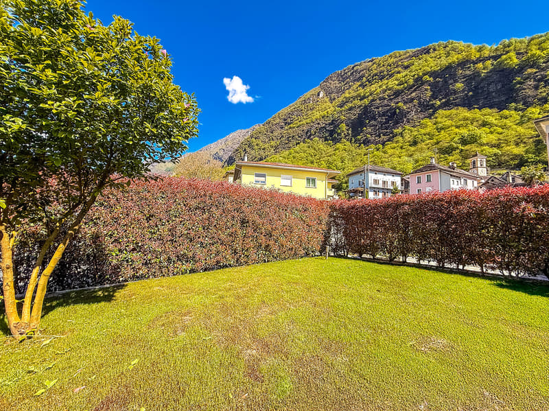 The image shows a residential area with colorful houses surrounded by lush greenery. There is a well-manicured lawn in the foreground, and the houses have balconies or terraces visible. The background features a mountainous landscape with dense vegetation