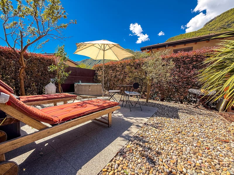 Cozy outdoor patio with wooden lounge chairs, a yellow umbrella, and a gravel path surrounded by lush greenery and plants.