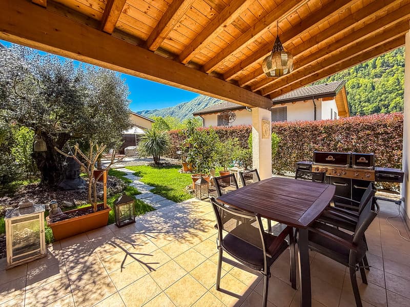 Covered patio with wooden beamed ceiling, outdoor dining table and chairs, barbecue grill, potted plants and decor, surrounded by lush greenery and mountains in the background