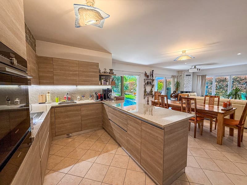 Spacious open-plan kitchen with wooden cabinets, granite countertops, tiled flooring, and a dishwasher. The kitchen leads into a dining area with a wooden table and chairs.