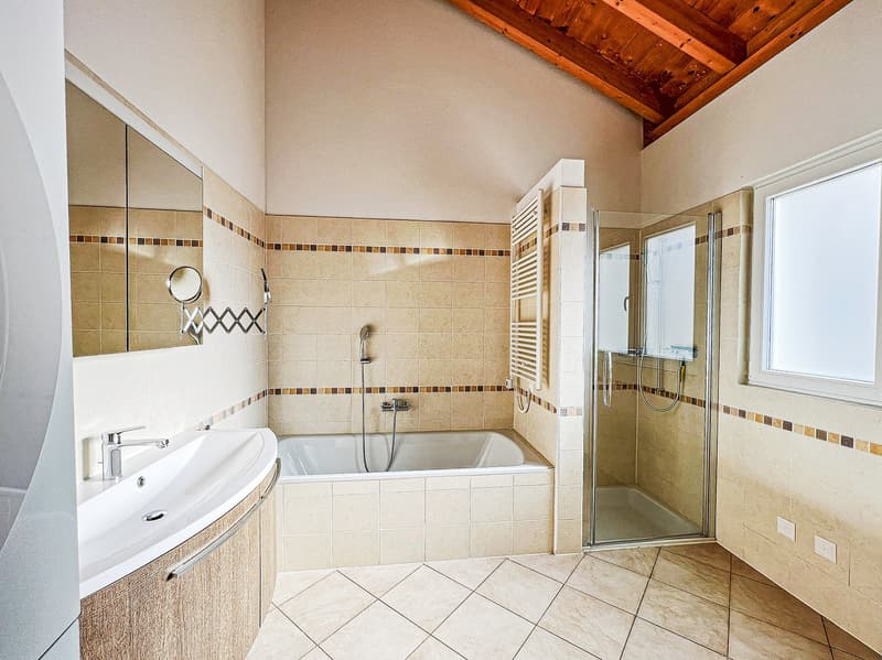 Spacious bathroom with tiled walls, a bathtub, a separate shower enclosure, and a vanity sink. The room has a vaulted ceiling with exposed wooden beams.