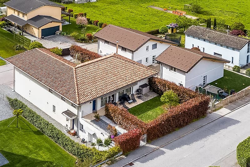 Two-story residential building with a red tile roof, surrounded by a well-maintained garden with lush greenery. The property also includes a garage and a parking space.