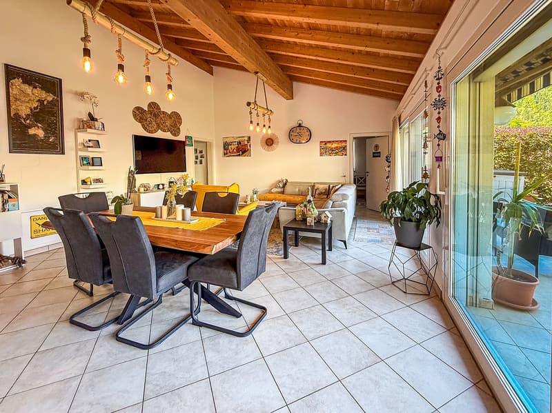Open-concept living and dining area with wooden beamed ceiling, tiled flooring, large windows, wooden dining table with chairs, sofa, and various decorative elements such as wall art and hanging lights.
