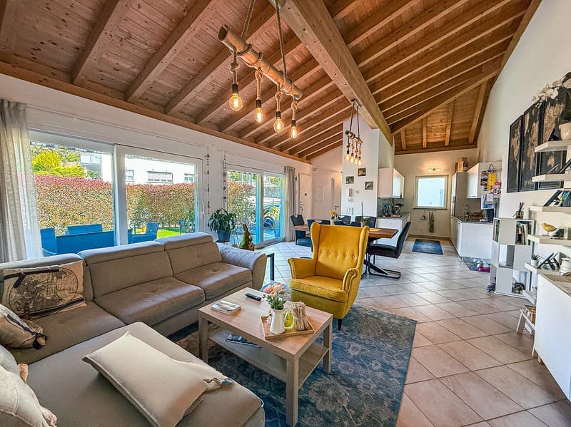 Open-concept living room with wooden beamed ceiling, large windows overlooking a balcony or terrace, brown sofa, yellow armchair, wooden coffee table, and various decor elements