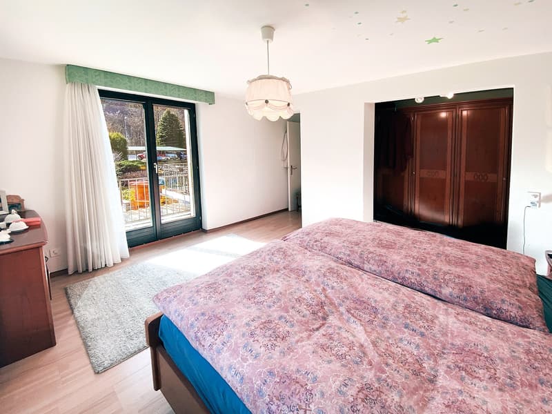 A bedroom with two twin beds, white curtains, a white rug, a chandelier, a wooden desk, a sliding door to a balcony, a cabinet, and a power outlet.
