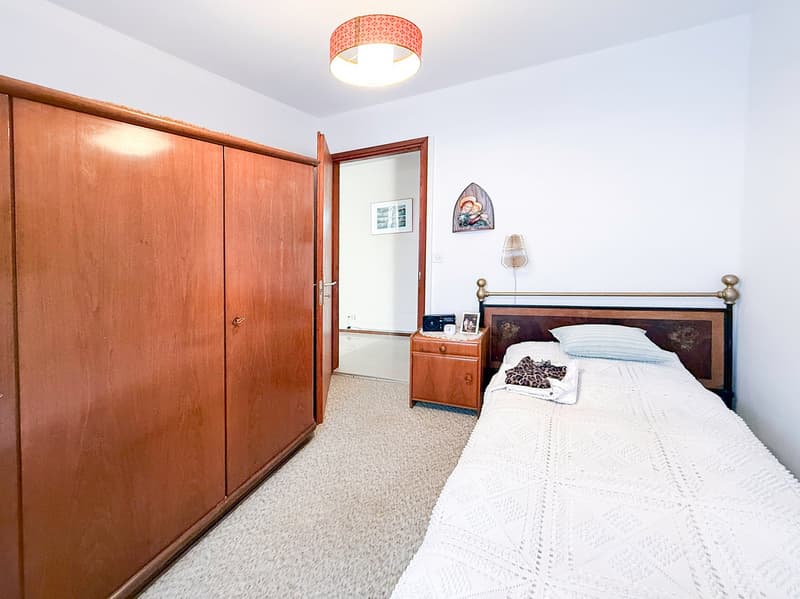 The image shows a bedroom with a wooden wardrobe, a wooden bed frame, a nightstand, and a patterned ceiling light fixture. The room has white walls and a carpeted floor.