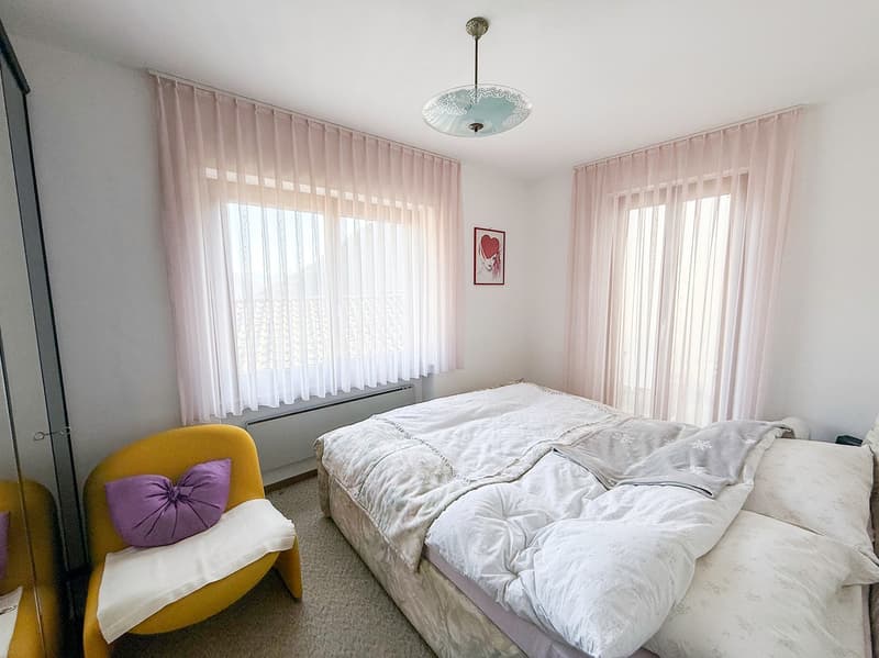 Spacious bedroom with white walls, large windows with sheer curtains, a queen-sized bed with white bedding, a yellow armchair with a purple pillow, and a decorative light fixture on the ceiling.