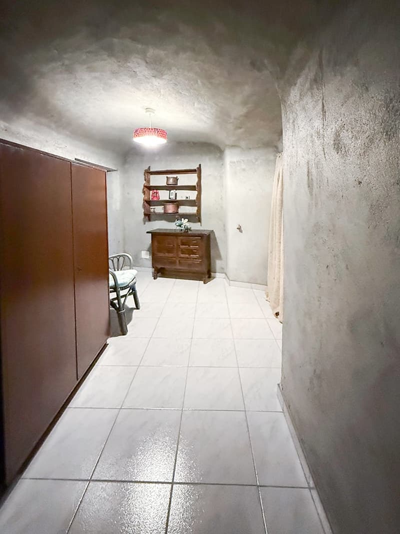 The image shows a hallway or entryway with white tile flooring, a wooden dresser or cabinet, and a red chandelier-style light fixture. The walls appear to be unfinished or in need of repair, with a rough, textured appearance. The overall atmosphere seems