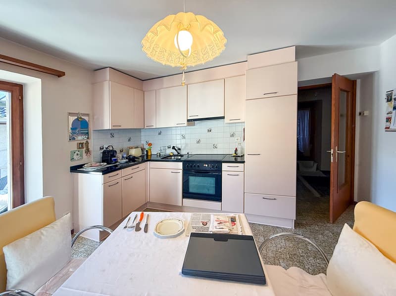 Fully equipped kitchen with white cabinets, black countertops, tiled backsplash, oven, and a decorative pendant light fixture. There is a dining table with chairs in the foreground.