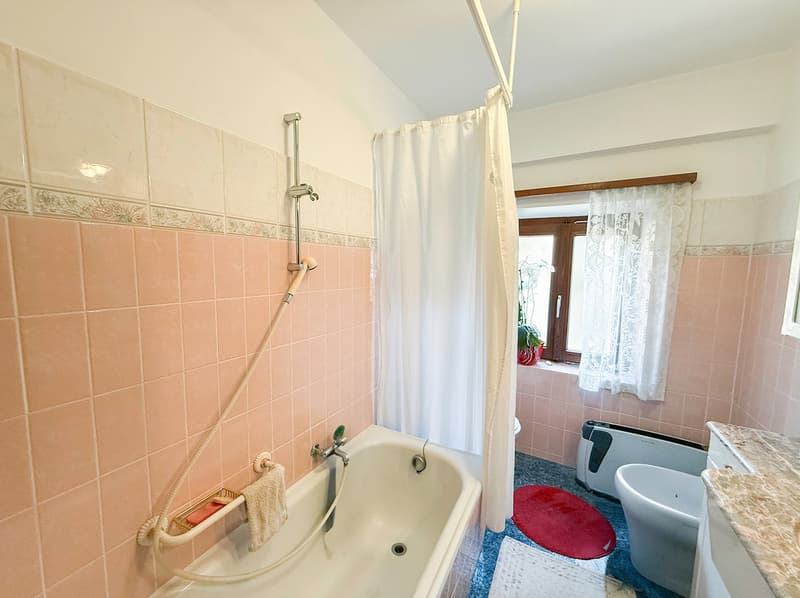 Bathroom with pink tile walls, a bathtub with a shower curtain, a toilet, and a window with lace curtains.