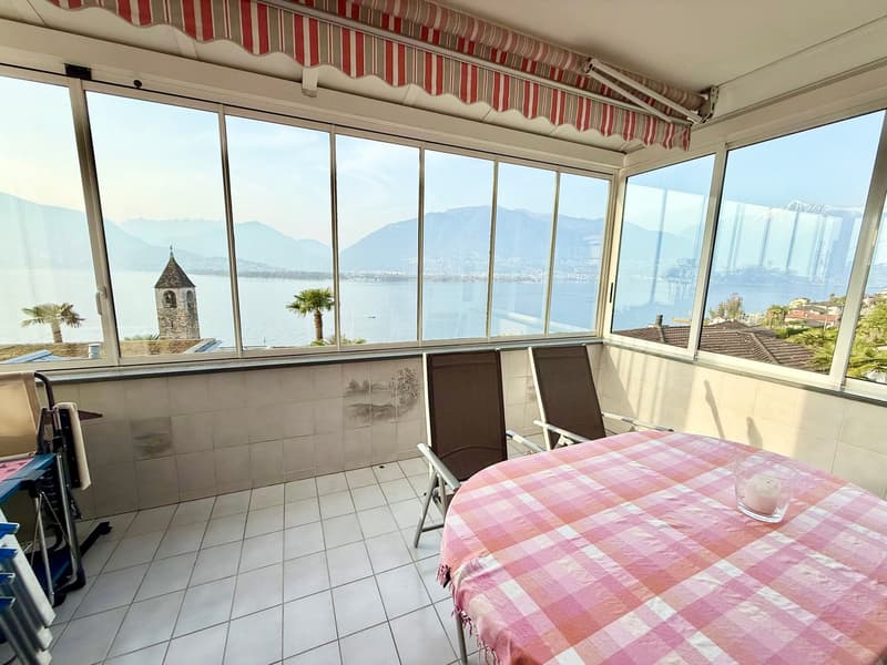 Balcony with large windows, tiled floor, table with pink and white checkered tablecloth, two chairs, view of the sea and mountains