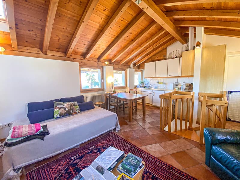 Open-plan living and dining area with a sofa, dining table, kitchen, and wooden beams on the ceiling.