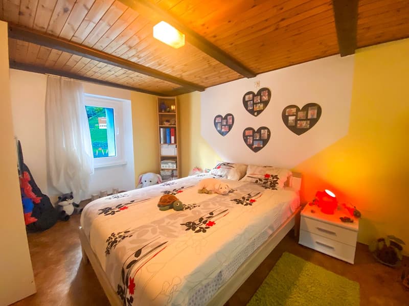 Bedroom with bed, nightstand, rug, wooden ceiling, yellow wall, window with curtains, shelf with books and toys, lamp, and plants.
