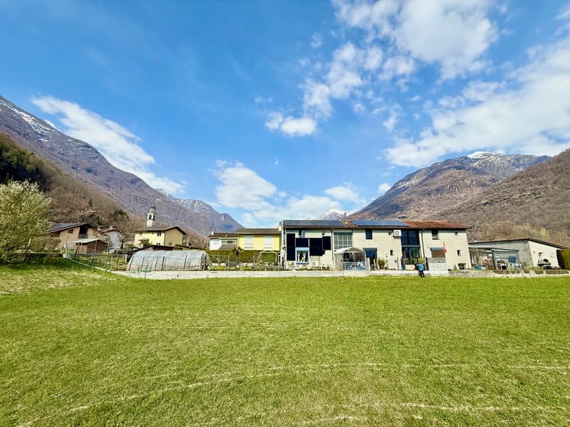 House with solar panels, surrounded by mountains, large green field, several buildings in the background