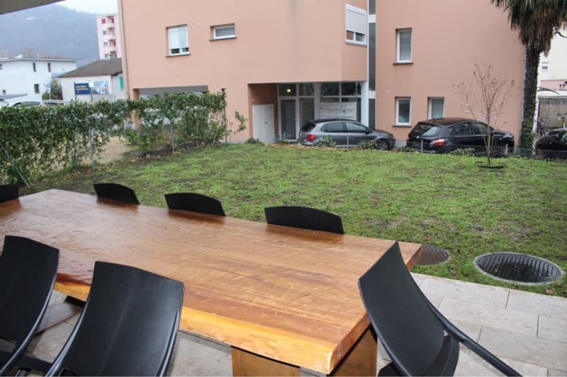 A garden with a wooden table and black chairs, a grassy area, and a building with parked cars in front.