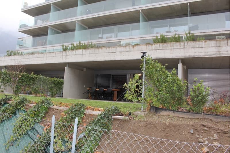 Modern apartment building with glass balconies, concrete structure, outdoor dining area, garden, and fenced yard