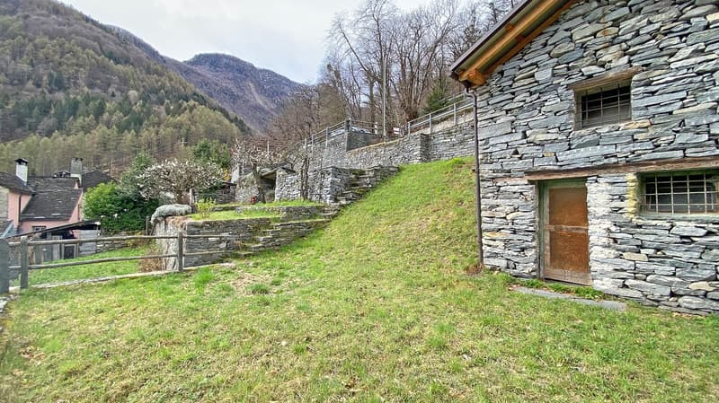 Stone house, raised ground floor, wooden door, stone walls, grassy slope, wooden fence, mountain view