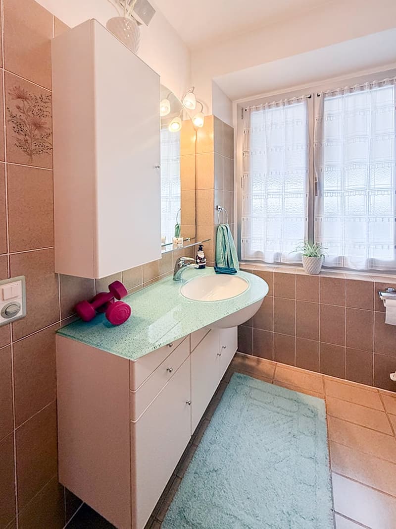 Bathroom: White cabinet, green countertop, white sink, white tiles, window with white curtain, small potted plant, towel and dumbbells