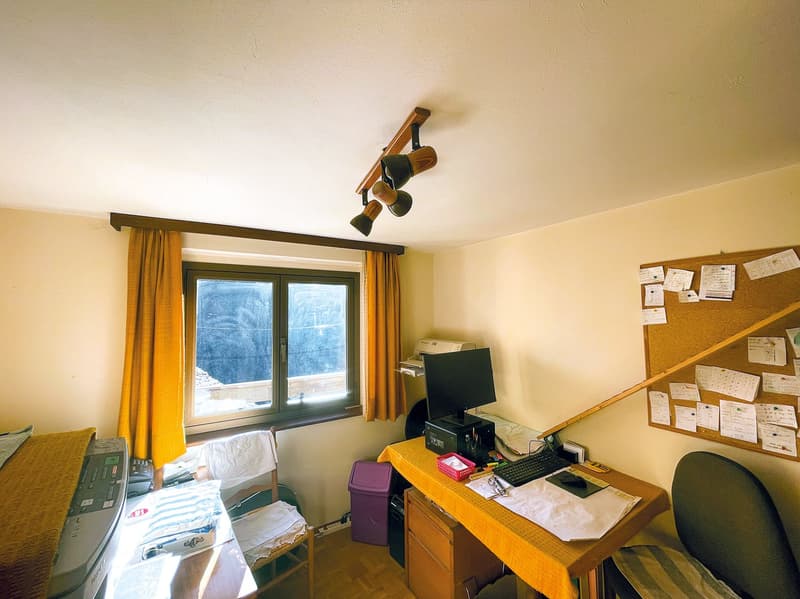 The image shows a bedroom with a desk, computer, and various office supplies. The room has a large window overlooking what appears to be a snowy outdoor scene. The walls are painted a light color, and the room has a cozy, lived-in feel.