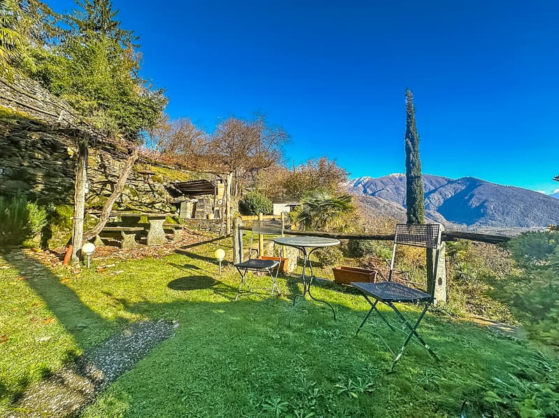 A garden with a table and chairs, surrounded by grass and plants, with a stone wall and a view of the mountains.