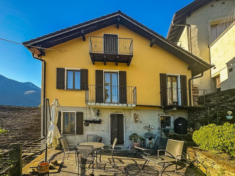 Two-story yellow house with a balcony, black shutters, and a patio area with outdoor furniture.
