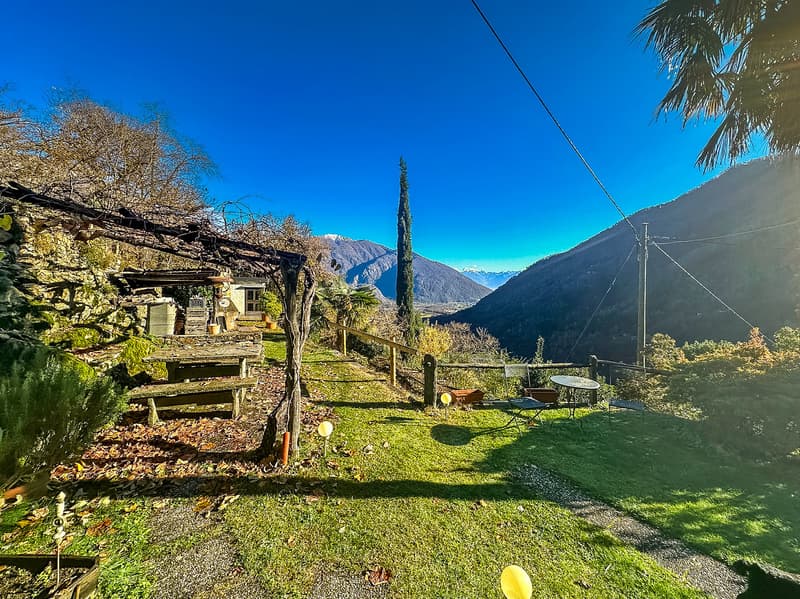 Outdoor area with a wooden table, chairs, and a bench, surrounded by greenery and a stone wall, with a view of mountains in the distance.