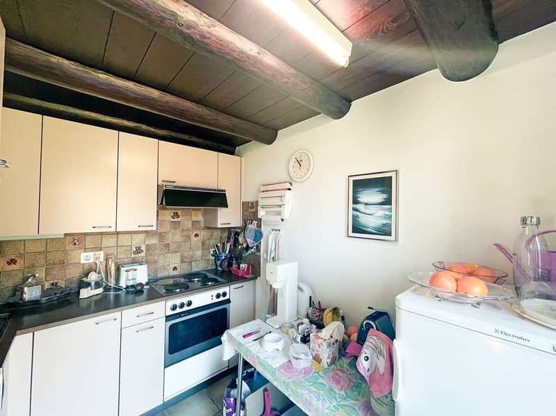 The image shows a kitchen with white cabinets, a tiled backsplash, and an electric stove. There is a dishwasher visible, as well as various kitchen appliances and decor items on the countertop. The ceiling has exposed wooden beams, adding a rustic touch t