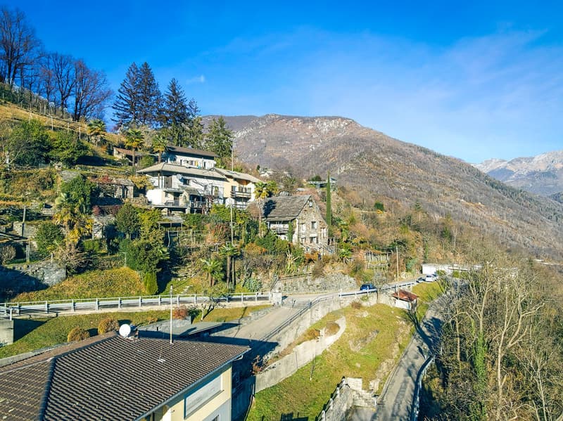 The image shows a picturesque mountain village nestled in a lush, green landscape. The buildings have balconies and terraces, and the surrounding area is dotted with trees and vegetation. In the background, there are tall, rugged mountains under a clear b