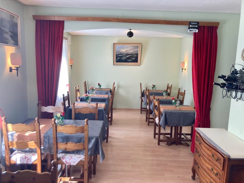 The image shows a cozy dining room with wooden furniture, including a long dining table with chairs, a wooden dresser, and red curtains. The walls are painted in a light green color, and there is a framed artwork hanging on the wall. The room has a warm a