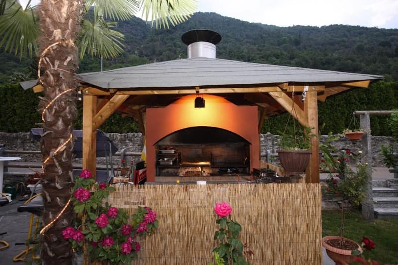 Wooden gazebo-style structure with a thatched roof, housing an outdoor pizza oven or fireplace. The structure is surrounded by potted plants and flowers, creating a cozy and rustic atmosphere.